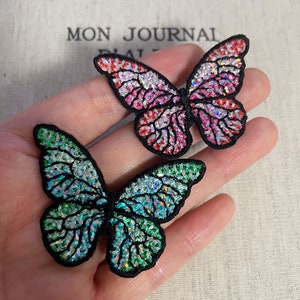 May include: Two embroidered butterfly patches. One butterfly has pink, red, and white wings, the other has green and blue wings. Both have black outlines and are embellished with iridescent sequins. The background has the text "MON JOURNAL".