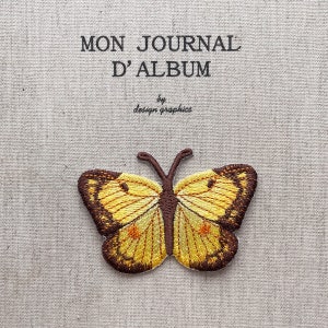 Può includere: Toppa a farfalla ricamata con ali gialle e marroni, dettagliata con cuciture intricate. La toppa è posta su un tessuto chiaro e strutturato con il testo "MON JOURNAL D'ALBUM" e "by design graphics".