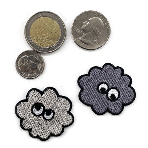 K&ouml;nnte beinhalten: Draufsicht auf drei M&uuml;nzen: ein Vierteldollar, ein Zehn-Cent-St&uuml;ck und eine Zwei-Euro-M&uuml;nze. Zwei gestickte Wolkenaufn&auml;her mit Wackelaugen sind ebenfalls zu sehen. Die Aufn&auml;her sind grau mit schwarzen Umrissen und wei&szlig;en und schwarzen Augen.