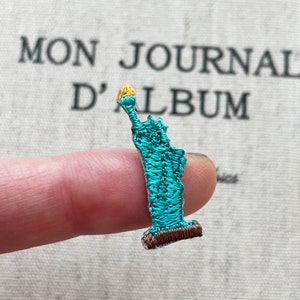 May include: A miniature embroidered Statue of Liberty patch in turquoise, with a brown base and a yellow torch. The patch is held between fingers, with the text "MON JOURNAL D'ALBUM" in the background.