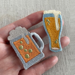 以下が含まれることがあります： ビールジョッキとグラスをあしらった刺繍パッチ。オレンジ、黄色、青のアクセントとシルバーグレーの縁取りが施されています。ビールグラスには泡のディテールがあります。衣類やアクセサリーに最適です。