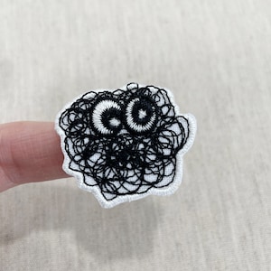 Peut inclure: Écusson brodé avec un design fantaisiste. Le patch a une bordure blanche et un motif noir griffonné avec deux grands yeux circulaires. Les yeux sont blancs avec des centres noirs, donnant au patch un aspect de dessin animé.