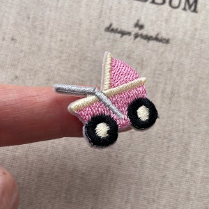 Op de afbeelding: Geborduurde roze kinderwagen patch met zwarte wielen en een zilveren handvat. De patch heeft een crèmekleurige rand en staat op een lichtbeige achtergrond. De patch is een schattig accessoire voor babyartikelen.