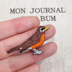 以下が含まれることがあります： オレンジ色の胸の羽、黄色のくちばし、白いアクセントが付いた茶色の鳥の刺繍パッチ。パッチは手で持たれており、背景には「MON JOURNAL ALBUM」の文字が見えます。