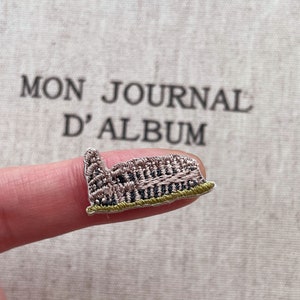 May include: Embroidered patch of a building with a brown and tan facade, a green base, and a small tower. The patch is held up by a finger. The text "MON JOURNAL D'ALBUM" is in the background.