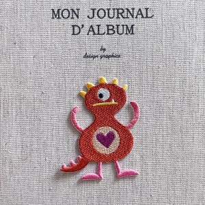 Könnte beinhalten: Ein Stoff-Journal mit dem Text "MON JOURNAL D'ALBUM" und einem roten Monster-Aufnäher. Der Aufnäher hat ein lila Herz, gelbe Stacheln und rosa Arme und Füße. Der Text "by design graphics" steht darunter.