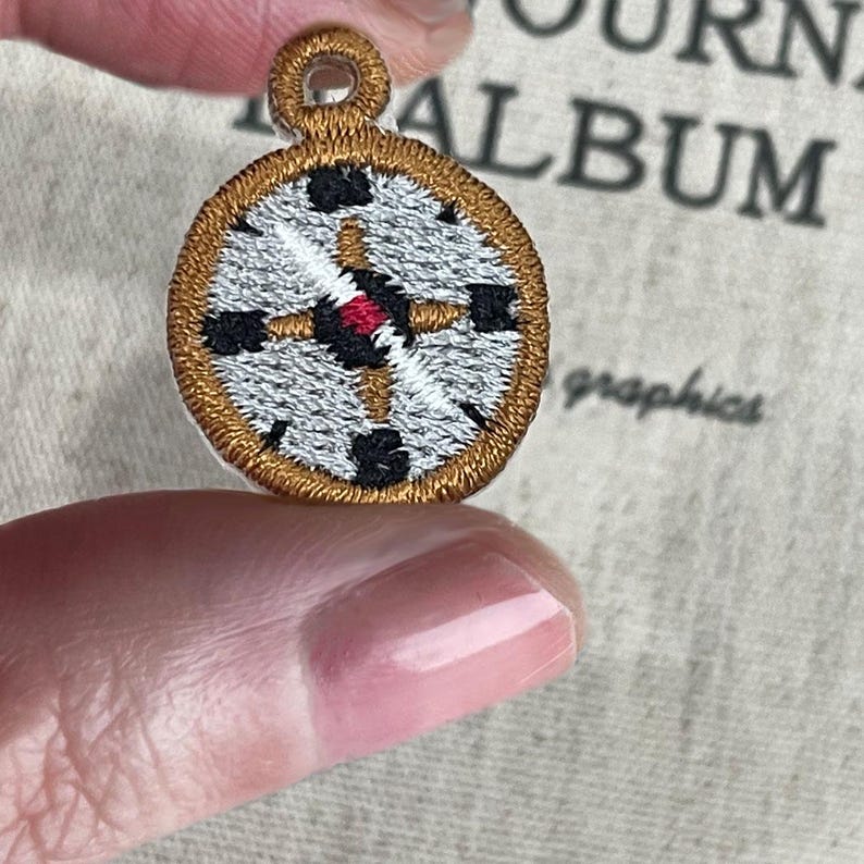 May include: Embroidered pendant with a compass design. The pendant has a gold border, a silver-grey centre, and black and red accents. It has a loop at the top for hanging. The background includes the words "JOURNAL ALBUM graphics."