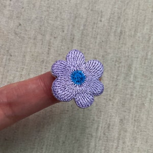 May include: Embroidered flower patch with a five-petal design. The petals are a light purple color, and the center is a bright blue. The patch is made with a detailed thread pattern.