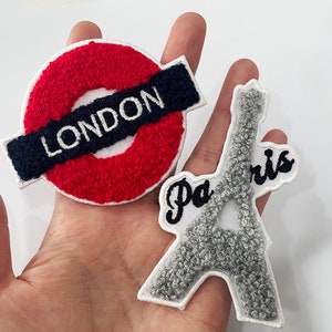 Könnte beinhalten: Zwei gestickte Aufnäher: Einer zeigt einen roten Kreis mit einem marineblauen Banner mit der Aufschrift "LONDON", der andere den Eiffelturm in Grau mit "Paris" in schwarzer Schrift. Beide Aufnäher haben ein strukturiertes, flauschiges Aussehen.