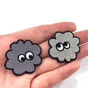 K&ouml;nnte beinhalten: Zwei gestickte Wolkenaufn&auml;her mit Wackelaugen. Einer ist grau, der andere hellgrau. Beide haben schwarze Umrisse und wei&szlig;e und schwarze Augen. Die Aufn&auml;her sind klein und wahrscheinlich f&uuml;r Kleidung oder Accessoires.