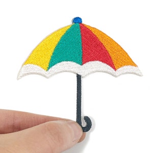 May include: Embroidered patch of a colorful umbrella with a yellow, green, and red canopy. The umbrella has a white trim and a dark gray handle. The patch is held by a hand against a white background.