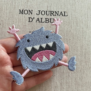 May include: Embroidered patch featuring a blue monster with a wide open mouth, white teeth, and a pink tongue. The monster has pink arms and legs. The background includes the text "MON JOURNAL D'ALBU".
