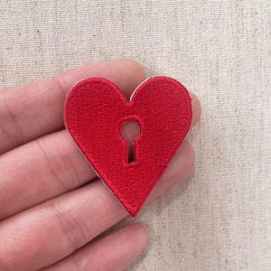 May include: A red heart-shaped embroidered patch with a keyhole design. The patch is made of red thread and has a white border. The keyhole is in the center of the heart.