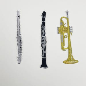 May include: Three embroidered patches featuring musical instruments: a silver flute, a black clarinet with silver keys, and a gold trumpet with silver accents. Each patch displays detailed stitching to represent the shape and components of the instruments.