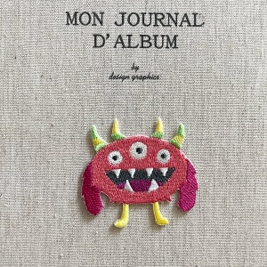 Könnte beinhalten: Gestickter rosa Monster-Aufnäher mit drei Augen, gelben Hörnern und einem breiten Grinsen. Der Aufnäher befindet sich auf einem beigefarbenen Stoffhintergrund mit dem Text "MON JOURNAL D'ALBUM" und "by design graphics".