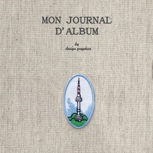 Może przedstawiać: Beżowa okładka dziennika z tkaniny z napisem "MON JOURNAL D'ALBUM" i "by design graphics". Na okładce znajduje się haftowana naszywka z wieżą z czerwonymi i białymi akcentami.