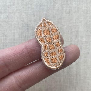 May include: Embroidered patch in the shape of a peanut. The patch features a tan border and a textured, orange and tan interior, resembling the shell of a peanut. The patch is held between two fingers.