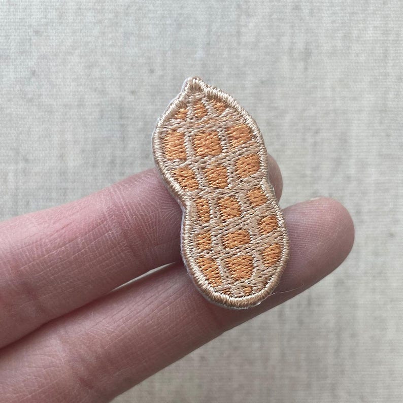 May include: Embroidered patch in the shape of a peanut. The patch features a tan border and a textured, orange and tan interior, resembling the shell of a peanut. The patch is held between two fingers.
