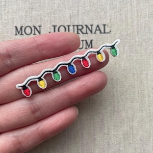 May include: An embroidered patch featuring a string of seven colorful Christmas lights. The lights are arranged in a wavy line and include red, yellow, blue, and green bulbs with a white border and black wire detailing on a white fabric base.