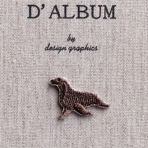 以下が含まれることがあります： 白の縁取りが付いた茶色の犬の刺繍パッチ。テクスチャ加工された淡いベージュ色の生地にあります。「D'ALBUM」の文字が黒色で、「by design graphics」の上にあります。