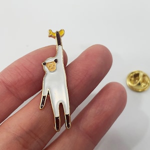 May include: A white and gold enamel pin depicting a cartoon monkey hanging from a branch. The monkey has a tan face and brown feet and hands. A gold pin back is also visible.