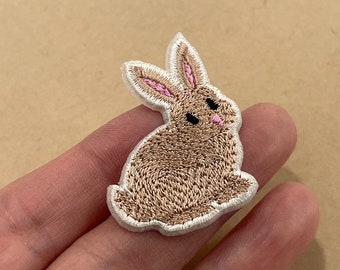 May include: Embroidered patch of a seated brown bunny with pink inner ears and a white border. The bunny has black eyes and a pink nose. This patch is suitable for clothing, bags, or other fabric items.