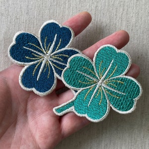 Puede incluir: Dos parches bordados con forma de trébol, uno turquesa y otro azul marino, con detalles dorados y blancos. Los parches se sostienen en una mano, mostrando la intrincada costura y el diseño de las aplicaciones florales.