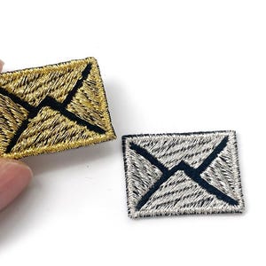 Könnte beinhalten: Zwei gestickte Aufnäher in Umschlagform. Einer ist goldfarben mit schwarzen Nähten, gehalten von einer Hand. Der andere ist silberfarben mit schwarzen Nähten. Beide sind rechteckig mit klassischem Umschlagdesign.