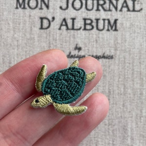 May include: Embroidered sea turtle patch with a dark green shell and gold-colored flippers and head. The patch is small and detailed, perfect for adding to clothing or accessories. The background has text that reads "MON JOURNAL D'ALBUM".