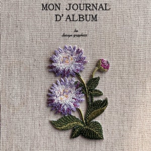 Könnte beinhalten: Bestickter Notizbuchumschlag mit dem Text "MON JOURNAL D'ALBUM" und "design graphics". Der Umschlag zeigt ein detailliertes Blumendesign mit lila, weißen und gelben Blüten, grünen Blättern und einer Knospe. Der Hintergrund ist ein strukturierter, beiger Stoff.