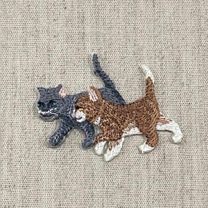 May include: Embroidered patch featuring two cats. One cat is gray, and the other is brown and white. The cats are depicted in a walking pose, with their tails raised. The patch is sewn onto a beige fabric.