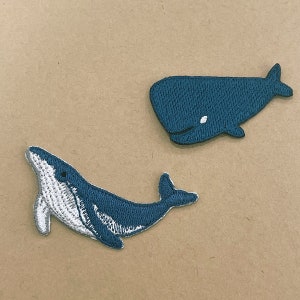May include: Two embroidered whale patches. One patch depicts a whale in profile, with a white belly and a dark blue body. The other patch shows a whale from above, in a dark blue color, with a white spot.