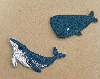 Op de afbeelding: Twee geborduurde walvispatches. Eén patch toont een walvis in profiel, met een witte buik en een donkerblauwe romp. De andere patch toont een walvis van bovenaf, in donkerblauwe kleur, met een witte vlek.