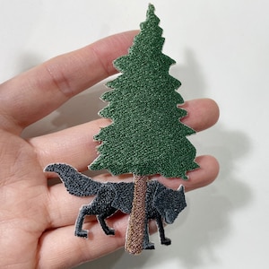 May include: Embroidered patch depicting a black wolf beneath a green pine tree. The patch has a detailed design with textured stitching, suitable for adding to clothing or accessories. The wolf and tree are set against a white background.