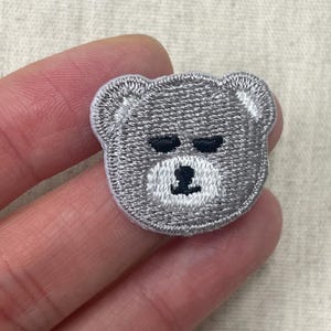 May include: A small, circular embroidered patch featuring a stylised bear face. The bear has light grey fur, two horizontal black lines for closed eyes, and a small black nose centred on a white muzzle area, all outlined with a white border.
