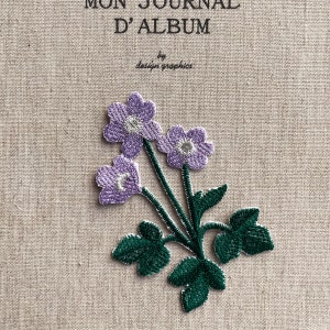 May include: Embroidered floral patch featuring three purple flowers with white centers and green leaves. The patch is on a beige fabric background. The text "MON JOURNAL D'ALBUM" and "by design graphics" are at the top.