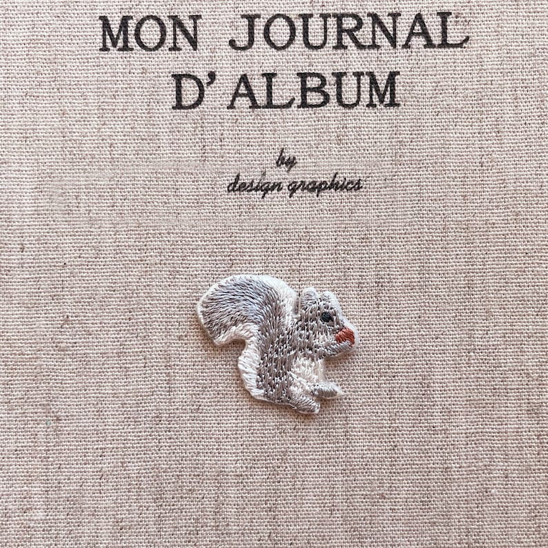 May include: Embroidered squirrel patch with gray and white fur, a red nose, and black eyes. The patch is on a beige fabric background with the text "MON JOURNAL D'ALBUM" and "by design graphics".