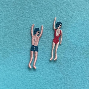 Può includere: Toppe ricamate di due nuotatori. Uno indossa pantaloncini blu, l'altro un costume da bagno rosso. Entrambi hanno le braccia alzate, indossando cuffie e occhialini. Lo sfondo è azzurro chiaro.