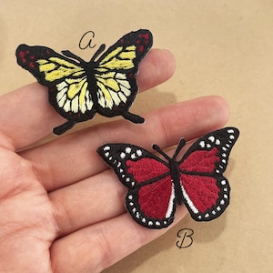 May include: Two embroidered butterfly patches. One butterfly has yellow and black wings with red accents. The other butterfly has red wings with white and black accents. Both patches are on a tan background.