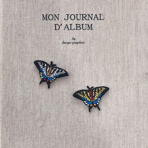 Pode incluir: Uma capa de diário com o texto "MON JOURNAL D'ALBUM" e "by design graphics". Dois patches de borboleta com detalhes em preto, azul, amarelo e branco são colocados na capa.