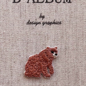 May include: A brown embroidered bear patch with a white border. The patch is on a beige fabric background. The text "D ALBUM by design graphics" is at the top of the image.