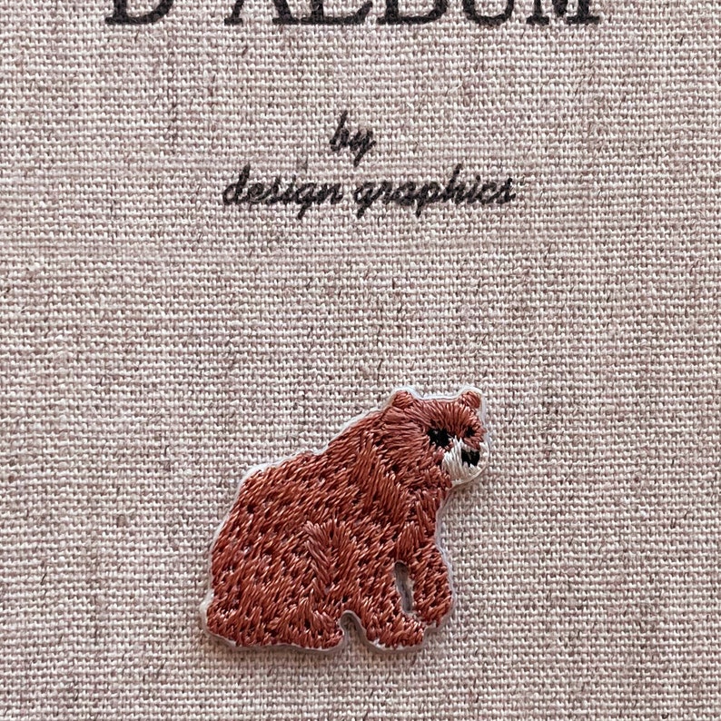 May include: A brown embroidered bear patch with a white border. The patch is on a beige fabric background. The text "D ALBUM by design graphics" is at the top of the image.