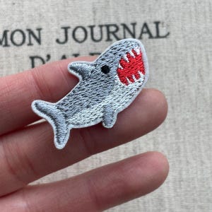 May include: Embroidered shark patch in shades of gray with a red mouth and white teeth. The patch is in the shape of a shark with a detailed design. The patch is small and appears to be made of fabric.