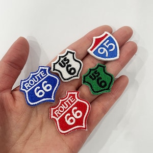 Puede incluir: Un conjunto de parches bordados con forma de señales de carretera estadounidenses. Los parches muestran los números 66 y 95, y el texto "US 6". Los colores incluyen azul, rojo, verde, blanco y negro.