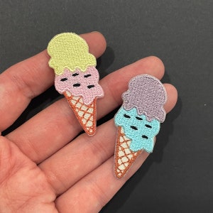 Peut inclure: Deux écussons brodés en forme de cornet de glace. L'un est jaune, rose et marron. L'autre est violet, bleu et marron. Chaque écusson a un motif de gaufre et trois boules de glace.