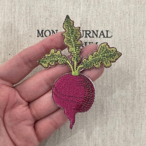 Puede incluir: Parche bordado con un rábano magenta vibrante con hojas verdes. El parche tiene un diseño detallado y un borde blanco. El fondo incluye el texto "MON JOURNAL".