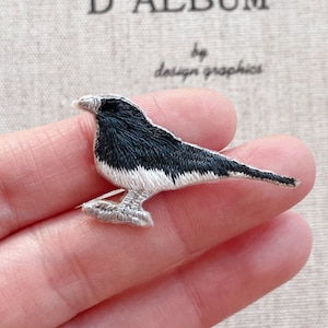 Puede incluir: Parche bordado de un pequeño pájaro con plumaje blanco y negro. El pájaro está representado de perfil, con detalles de costura finos. El texto "by design graphics" es visible. Ideal para coser.