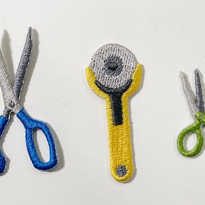Puede incluir: Parches bordados con herramientas de costura: unas tijeras con mango azul, un cortador rotatorio amarillo y unas tijeras con mango verde. Los parches tienen costuras detalladas sobre un fondo blanco.