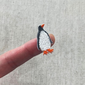 May include: Embroidered penguin patch with a black head and back, white chest, and orange feet and beak. The patch is small and detailed, perfect for adding to clothing or accessories.