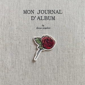 May include: Embroidered patch featuring a red rose with green leaves and a white border. The patch is on a textured, light gray background with the text "MON JOURNAL D'ALBUM" and "by design graphics".